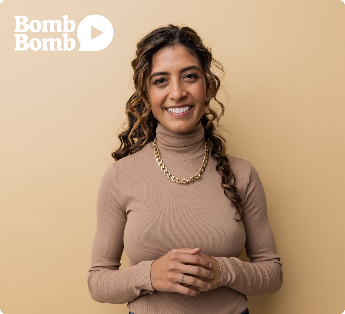 A smiling person stands confidently, wearing a beige turtleneck and a gold chain necklace. Behind them is the BombBomb logo, suggesting their affiliation with the brand, aiming to highlight the personal and professional connection that BombBomb video messaging seeks to foster.