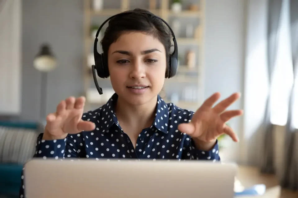 A woman with headphones gesturing while looking at a laptop screen.
