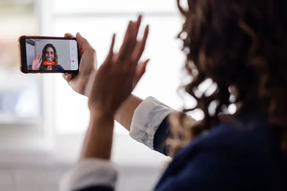 A person is recording a video call with another individual waving from the smartphone screen, likely engaging in a virtual interaction.